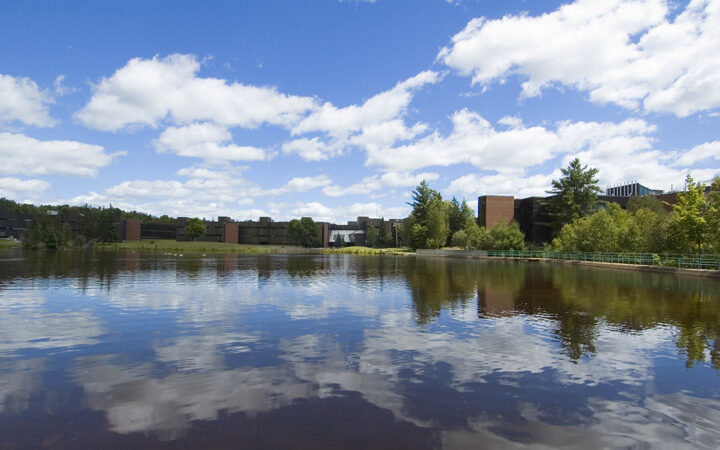 A pond reflecting clouds and trees, with Nipissing University buildings on the far shore