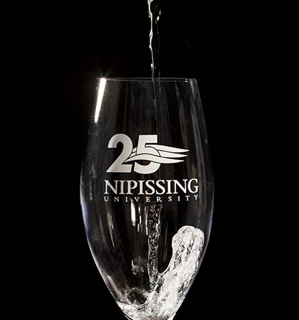Water being poured into a wine glass etched with the Nipissing University 25th anniversary logo, against a black background