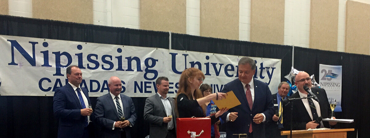 A ceremony on stage under a 'Nipissing University' banner, with officials standing at a podium and a table with a 25th anniversary cloth and a red box