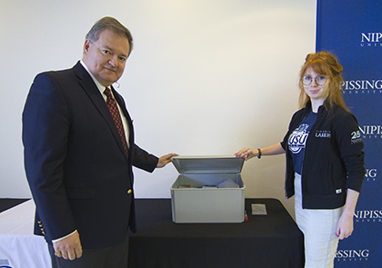 A man in a suit and a woman in a Nipissing University sweatshirt stand on either side of a table holding an open metal box