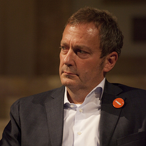 Close-up portrait of a serious man in a dark blazer with an orange button on the lapel