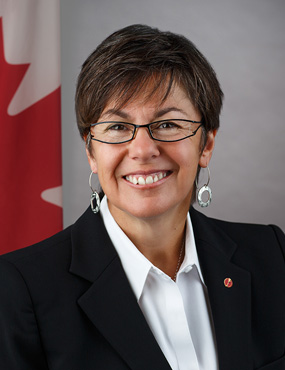 Portrait of a smiling woman with short brown hair and glasses, wearing a black blazer in front of a Canadian flag