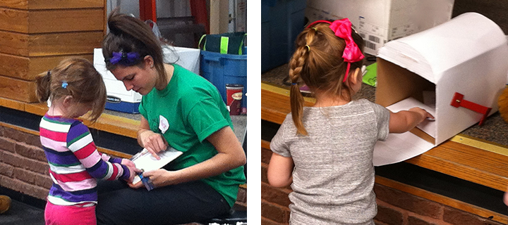 A young woman in a green shirt helping a small girl write, next to a second girl posting a letter in a cardboard mailbox