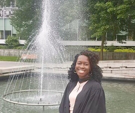 A smiling woman in a black graduation gown standing in front of an outdoor fountain