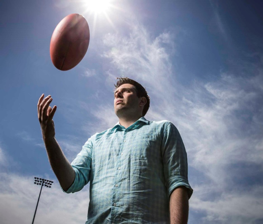 A man in a light blue checked shirt tosses a rugby ball into the air, photographed from below against a blue sky