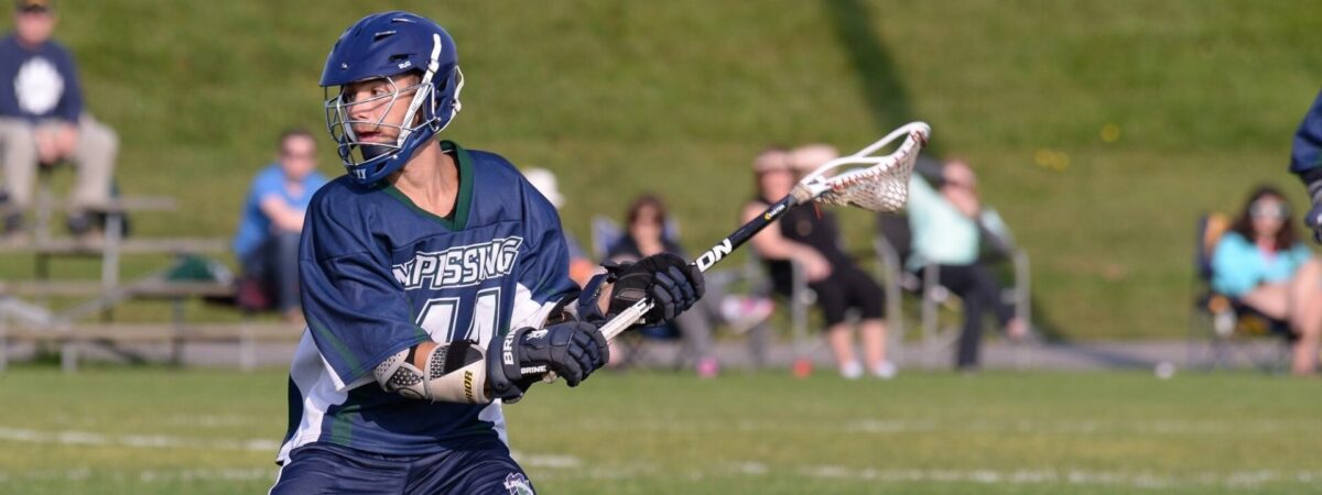 A Nipissing men's lacrosse player in a navy jersey cradles the ball in his stick on a grass field