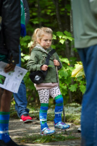 A young girl in a green jacket and colourful socks standing on a forest path holding a trail map