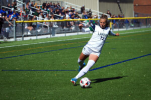 A Nipissing Lakers women's soccer player in a white number 12 jersey kicks the ball on a turf field
