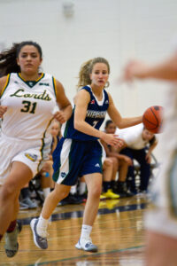 A Nipissing women's basketball player in navy dribbles the ball up the court with a defender in a white 'Lords' jersey close behind