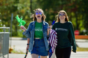 Two smiling students in Nipissing Lakers clothing walk outdoors, one waving a green pom-pom