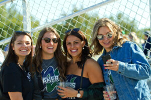 Four smiling friends in Lakers shirts and sunglasses pose behind a chain-link fence