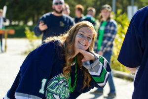 A smiling woman in a Nipissing Lakers jersey and green beads leans toward the camera outdoors with blue face gems