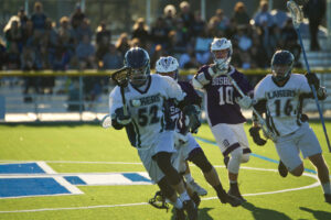 Nipissing Lakers lacrosse players in white jerseys run with the ball against players in purple on a turf field