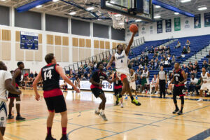 A Nipissing Lakers basketball player in white leaps to the hoop for a layup while defenders in black Canadore jerseys look on