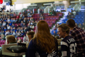 Spectators in Lakers jerseys sitting at a table in a stadium suite, with a crowd in the stands behind them