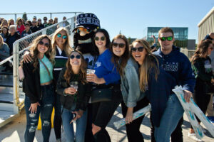 A group of smiling students in Nipissing Lakers clothing, including a mascot, pose together in front of the stands holding inflatable cheering sticks