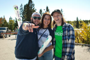 Three smiling people in Lakers clothing outdoors, one pointing at the camera, with an inflatable cheering stick in the background