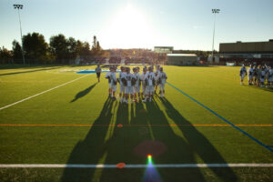Lacrosse players huddled on a turf field at sunset, casting long shadows