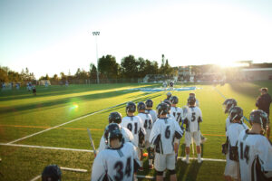 Nipissing Lakers lacrosse players in white jerseys stand on a turf field in low evening sun