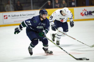 A Nipissing hockey player in a navy number 77 jersey skates with the puck as an opponent in white chases him