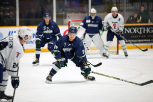 Nipissing hockey players in navy jerseys crouch ready on the ice in front of the net as opponents in white look on