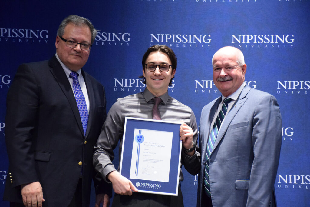 A young man in glasses holding a framed Dave Marshall Leadership Award certificate between two men in suits in front of a Nipissing University backdrop