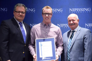 A smiling young man in a checked shirt holding a framed Dave Marshall Leadership Award certificate between two men in suits in front of a Nipissing University backdrop