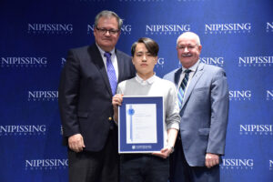 A young man in a grey sweater holding a framed Dave Marshall Leadership Award certificate between two men in suits in front of a Nipissing University backdrop