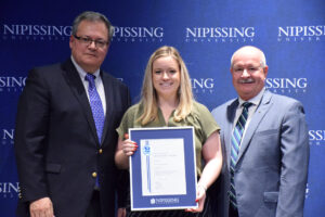 A smiling woman with long blonde hair holding a framed Dave Marshall Leadership Award certificate between two men in suits in front of a Nipissing University backdrop