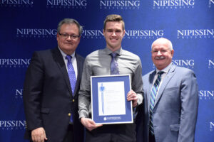 A tall young man in a grey shirt holding a framed Dave Marshall Leadership Award certificate between two men in suits in front of a Nipissing University backdrop