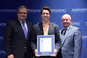 A young man in a grey blazer holding a framed Dave Marshall Leadership Award certificate between two men in suits in front of a Nipissing University backdrop