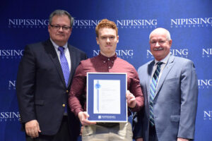 A red-haired young man in a burgundy shirt holding a framed Dave Marshall Leadership Award certificate between two men in suits in front of a Nipissing University backdrop