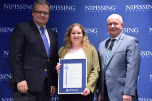 A young woman with curly hair and a green cardigan holding a framed Dave Marshall Leadership Award certificate between two men in suits in front of a Nipissing University backdrop