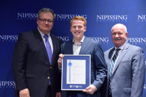 A smiling red-haired young man in a blue blazer holding a framed Dave Marshall Leadership Award certificate between two men in suits in front of a Nipissing University backdrop