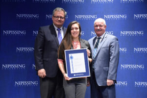 A young woman with long dark hair holding a framed Dave Marshall Leadership Award certificate between two men in suits in front of a Nipissing University backdrop