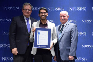 A young man in glasses and a cream blazer holding a framed Dave Marshall Leadership Award certificate between two men in suits in front of a Nipissing University backdrop