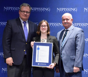 A young woman with glasses holding a framed Dave Marshall Leadership Award certificate between two men in suits in front of a Nipissing University backdrop