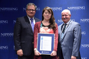 A young woman in a red cardigan holding a framed Dave Marshall Leadership Award certificate between two men in suits in front of a Nipissing University backdrop