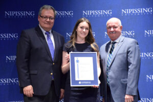 Two men in suits stand on either side of a young woman holding a framed Dave Marshall Leadership Award certificate in front of a Nipissing University backdrop