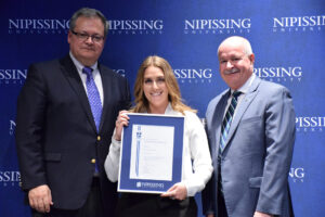 A smiling woman in a white blouse holding a framed Dave Marshall Leadership Award certificate between two men in suits in front of a Nipissing University backdrop