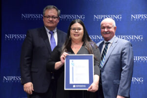 A woman with dark hair and glasses holding a framed Dave Marshall Leadership Award certificate between two men in suits in front of a Nipissing University backdrop