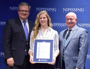 A young woman with long wavy hair in a striped blouse holding a framed Dave Marshall Leadership Award certificate between two men in suits in front of a Nipissing University backdrop