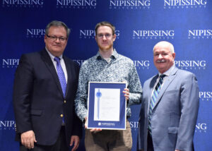 A young man with glasses in a patterned shirt holding a framed Dave Marshall Leadership Award certificate between two men in suits in front of a Nipissing University backdrop