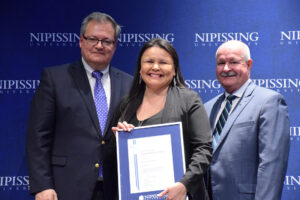 A woman smiling and holding a framed Dave Marshall Leadership Award certificate, standing between two men in suits in front of a Nipissing University backdrop