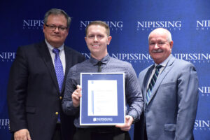 A smiling young man in a dark patterned shirt holding a framed Dave Marshall Leadership Award certificate between two men in suits in front of a Nipissing University backdrop