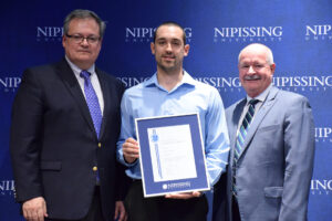 A man in a light blue shirt holding a framed Dave Marshall Leadership Award certificate between two men in suits in front of a Nipissing University backdrop