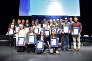 A group of about twenty award recipients standing and kneeling on a stage, each holding a framed certificate, under a screen reading 'Leadership Awards'