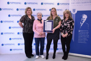 Four women pose in front of a Nipissing University backdrop and Dave Marshall Leadership Awards banner; one holds a framed certificate