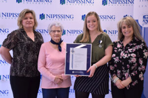 Four women pose close together in front of a Nipissing University backdrop; one holds a framed Dave Marshall Leadership Award certificate