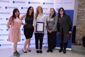 Five women pose in front of a Nipissing University backdrop and Dave Marshall Leadership Awards banner; one holds a framed certificate